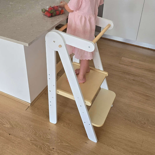 A toddler standing on the Four Little Monkeys White Adjustable Foldable Learning Tower at a kitchen surface.