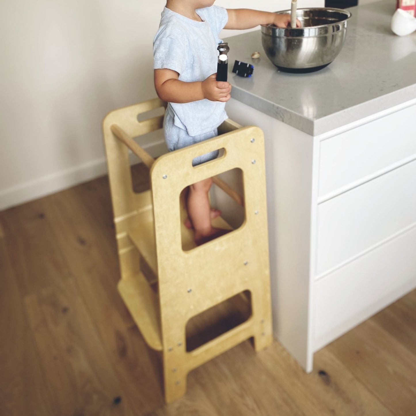 Child standing on 'Adjustable Learning Tower in Natural finish' by Four Little Monkeys, engaging in kitchen activities.