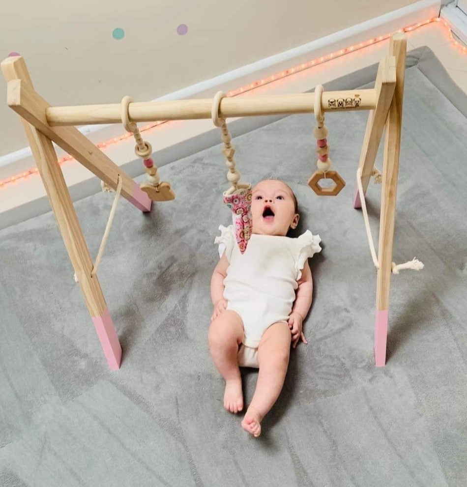 Adorable baby interacting and playing with the 'Baby Play Gym' in pink color by Four Little Monkeys showcasing its modern design with a splash of pink and three hanging toys.
