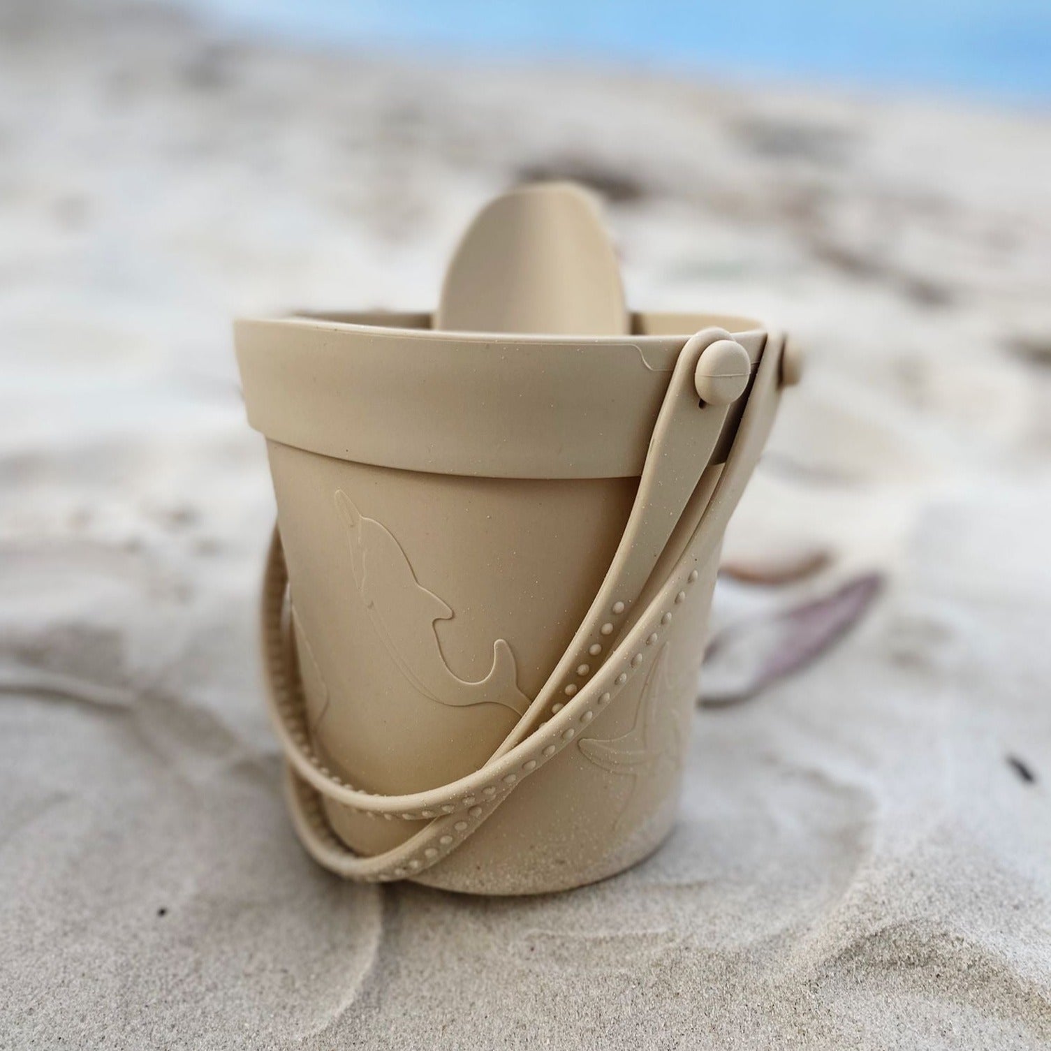 Sand Silicone Beach Bucket from the Silicone Beach Set is pictured on the sandy shore, demonstrating the durable and easy-to-clean nature of the beach toys in a real-life beach setting with natural seaside elements in the backdrop
