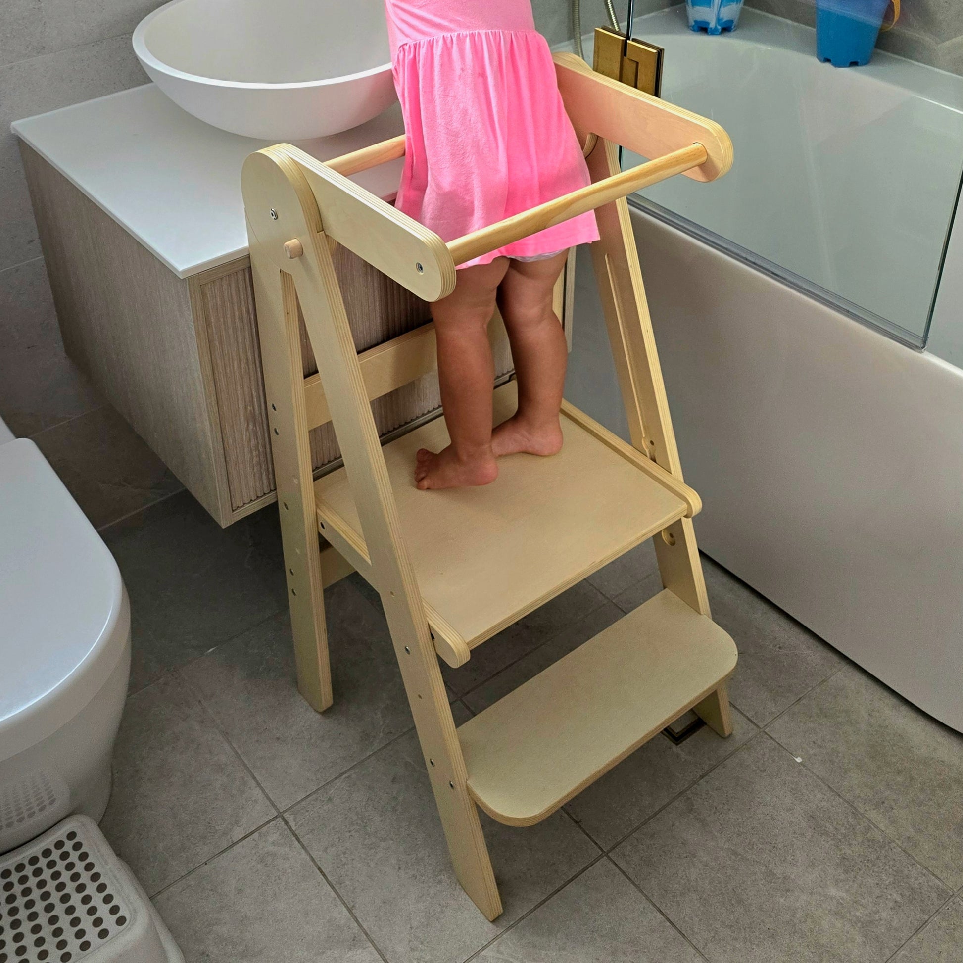 Toddler standing on Four Little Monkeys Natural Adjustable Foldable Learning Tower, washing hands at a bathroom sink.